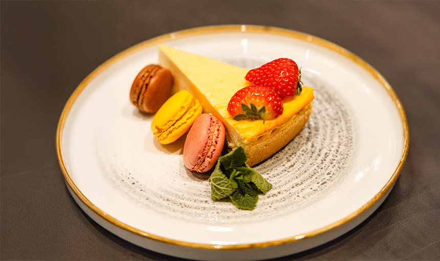 Käsekuchen mit pastellfarbenen Macarons auf einem eleganten weißen Teller, dekoriert mit zarten Streublumen und einem dezenten Puderzuckerhauch. Die Komposition zeigt eine raffinierte Dessert-Kreation mit cremigem Käsekuchengrund und luftigen französischen Mandelgebäck in sanften Pastelltönen. Frisch von unserem Restaurant für Sie als Dessert auf dem Teller.