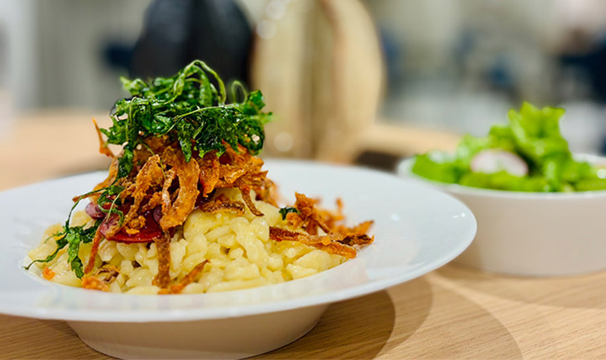 Das Bild zeigt einen Teller mit traditionellen Spätzle, modern interpretiert. Die goldgelben, buttrigen Spätzle sind mit frisch geriebenem Käse verfeinert, was für eine cremige und geschmackvolle Textur sorgt. Frische Kräuter als Garnitur verleihen dem Gericht eine aromatische Note.