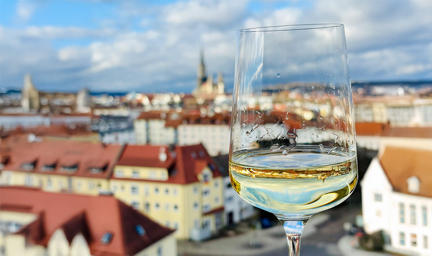 Das Bild zeigt eine atemberaubende Aussicht über die Dächer von Neu-Ulm von der Rooftop-Terasse des Heinrichs 161, während der Blick auf ein Glas Wein gerichtet ist.